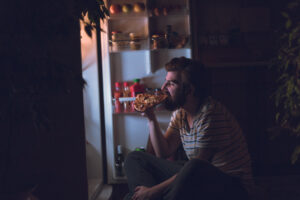 Een man eet ’s nachts een pizza op, zittend voor de ijskast.